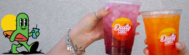 Cactus mascot beside two iced Dusty Coast Sodas (red and orange) against a gray wall.